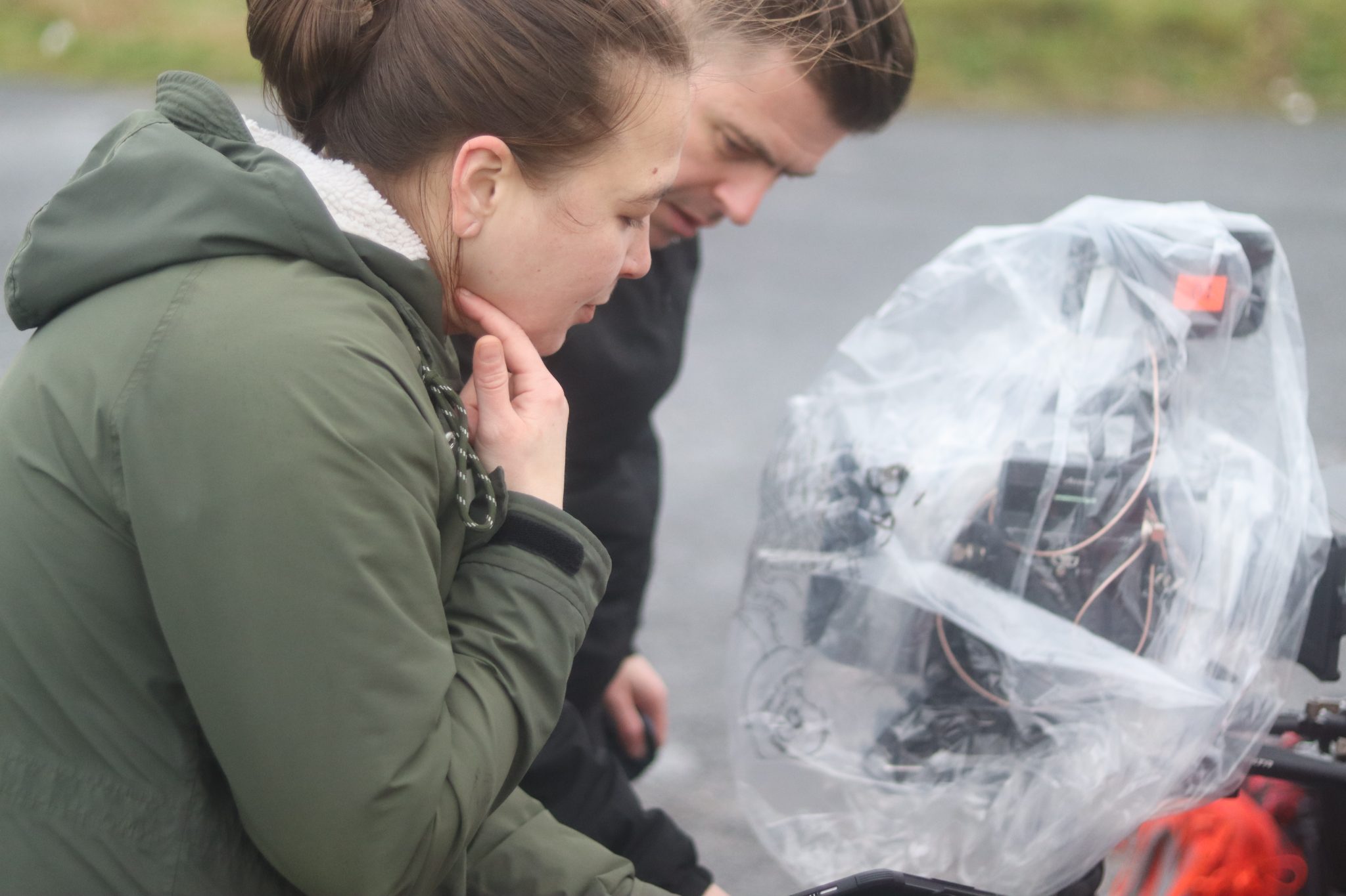Medium Shot of Klarrisa Webster and Will Clark looking at a camera that is protected from the rain with some plastic bag. Both Klarrisa and WIll are wearing rain jackets.